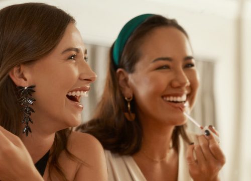 Photo of two happy women looking in a mirror, applying makeup