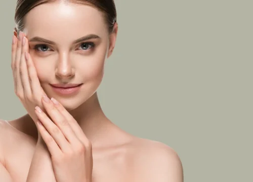 Studio portrait of a woman with bare shoulders and hands resting on her face against a neutral background.