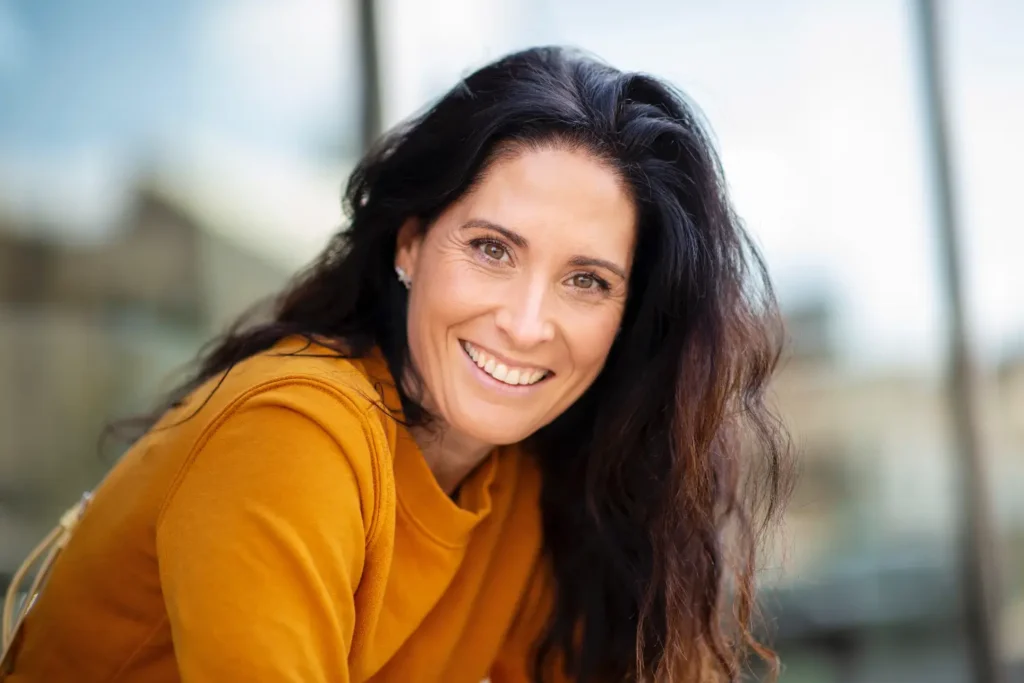 smiling woman with long dark hair wearing a mustard sweater outdoors