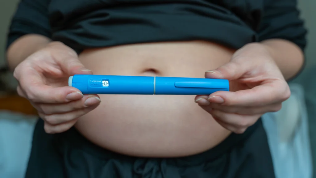 Hands holding blue injection pen in front of bare stomach
