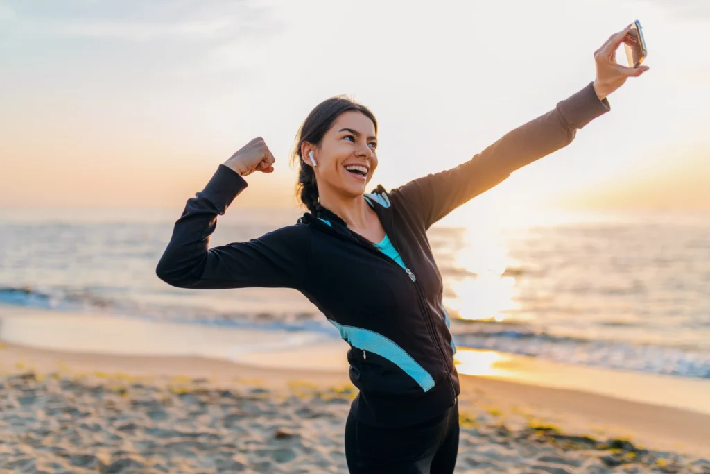Woman in athletic wear taking selfie on beach while flexing arm at sunrise