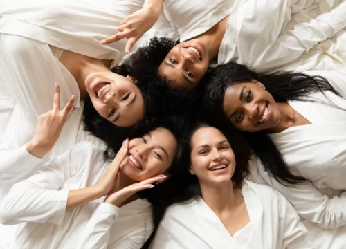 Group of women lying together on white bedding smiling at camera