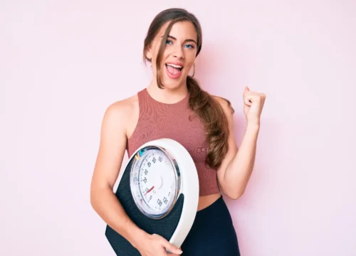 Woman holding a scale and raising her fist while smiling against a light background.