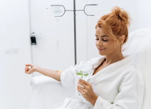 Person in a white robe seated during IV therapy while holding a glass of water.