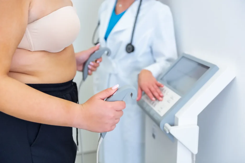 Woman holds body composition sensors while a medical professional operates a nearby assessment device.