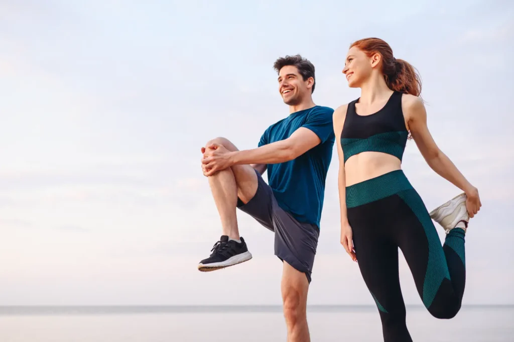 Man and woman in athletic clothing stretch their legs together outdoors near the water.