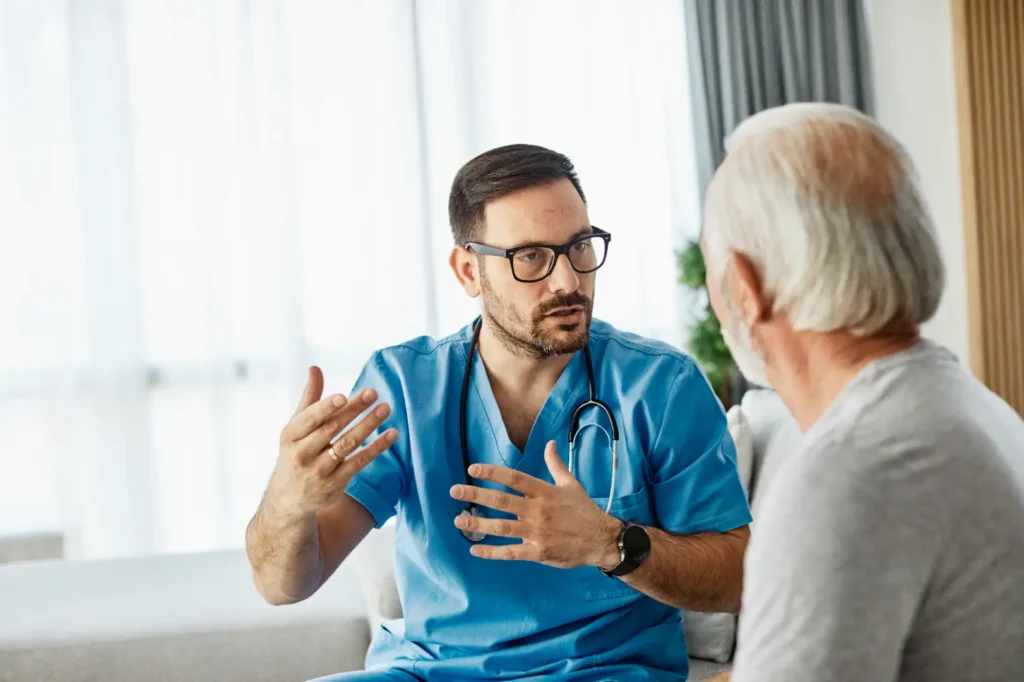 Doctor in blue scrubs gestures while speaking with an older man seated across from him.