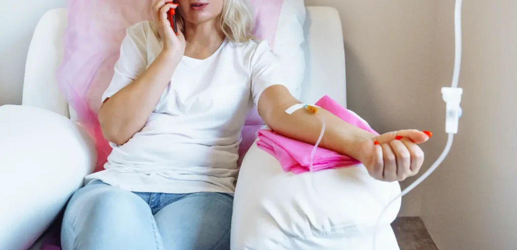 Person seated in a white chair with IV tubing attached to one arm and a phone held near their face.