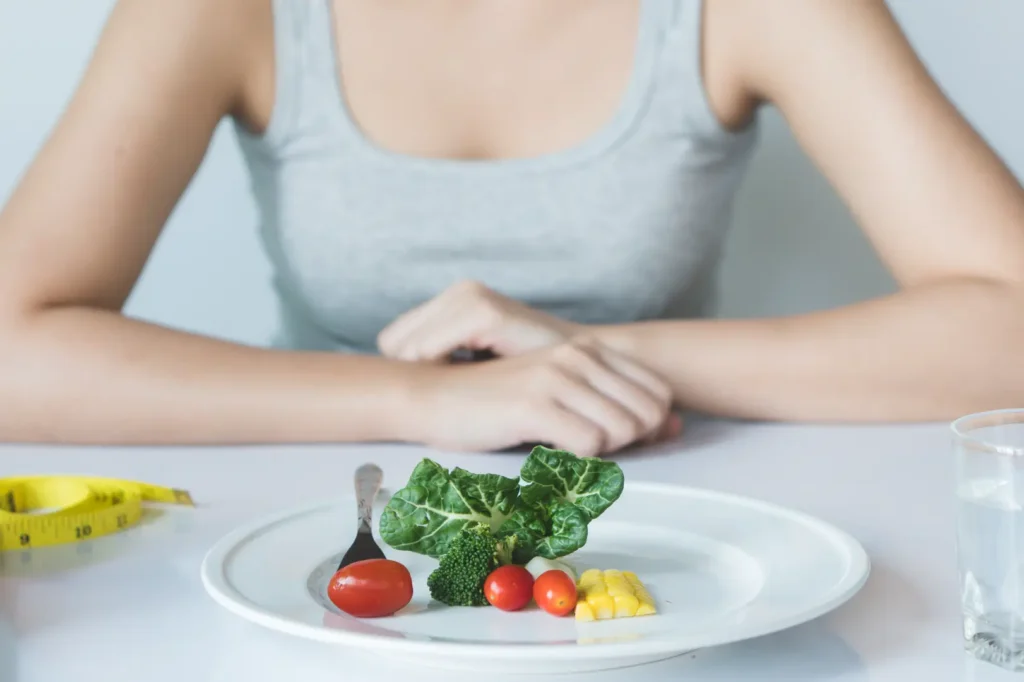 Small serving of vegetables on a white plate with a measuring tape and glass of water nearby.