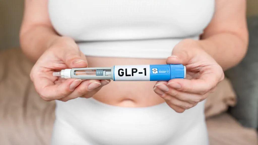 Woman holds an injection pen labeled GLP-1 horizontally in both hands in front of her abdomen.