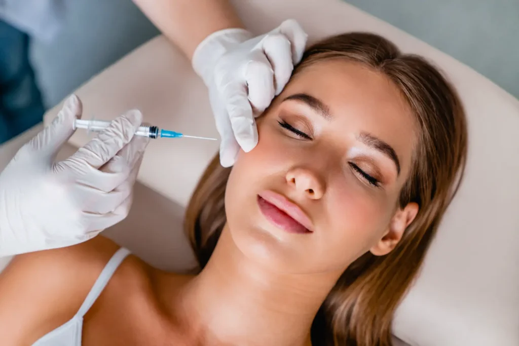 Woman lies with eyes closed while gloved hands position a syringe beside her cheek.