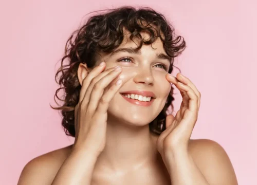 Smiling woman with curly hair touching both cheeks against a plain pink background.